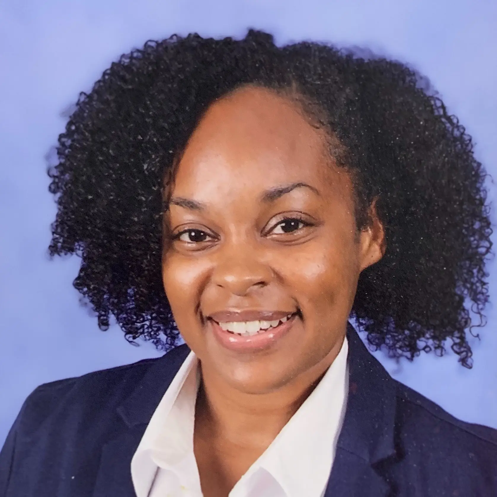 Portrait of a smiling woman with natural curly hair, wearing a navy blazer and white shirt, against a purple backdrop.