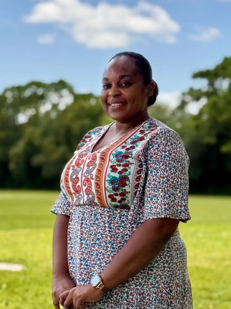 Smiling woman in a patterned dress standing in a sunny outdoor field with trees in the background.